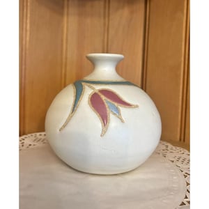 May include: A round, off-white ceramic vase with a narrow neck. The vase is decorated with a stylized floral design in pink, blue, and gold. The vase is sitting on a white doily.