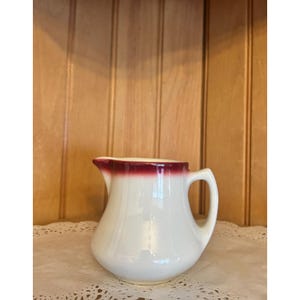 May include: White ceramic creamer with a burgundy rim. The creamer has a rounded body and a handle.