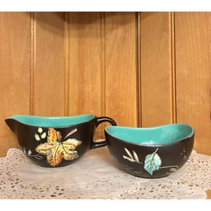 Puede incluir: Un juego de jarra de crema y azucarero de color marrón con interior azul turquesa y diseño de hojas. Las hojas están pintadas en tonos marrones, amarillos y verdes.
