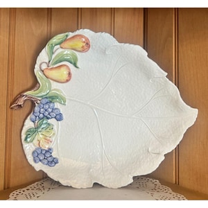 May include: White ceramic leaf-shaped serving platter with a textured surface. The platter is decorated with a branch of pears and grapes in shades of yellow, green, and blue. The edge of the platter is scalloped.