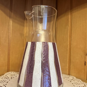 May include: A clear glass pitcher with a spout. The bottom half of the pitcher is decorated with alternating vertical stripes of white and dark purple. The pitcher is sitting on a white doily.
