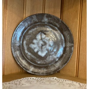 Puede incluir: Un plato de cerámica hecho a mano con un esmalte marrón oscuro y un diseño floral blanco en el centro. El plato tiene un borde ligeramente elevado y una apariencia rústica y texturizada. El plato tiene aproximadamente 25 cm de diámetro.