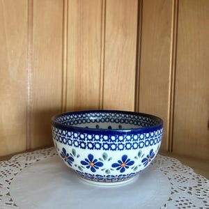 May include: A ceramic bowl with a white base and a blue floral pattern. The rim is dark blue with a geometric design. The bowl features blue and orange flower motifs. The bowl is sitting on a white doily.