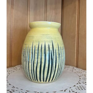 May include: A handmade ceramic vase with a light yellow glaze and vertical black lines. The vase has a rounded body and a slightly flared rim. It sits on a white doily.