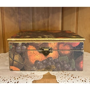 May include: A wooden box with a gold trim and a brass latch. The box is decorated with a vintage-style print of fruit, including grapes, apples, and oranges.