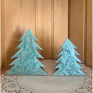 May include: Two turquoise-colored, rustic-style Christmas tree decorations. The trees have a textured surface and are displayed on a white, lacy doily. The background is a wooden wall.