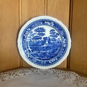 May include: A round ceramic plate featuring a blue and white transferware pattern. The central design depicts a landscape with a stone bridge, trees, and a building. The rim has a scalloped edge with a floral and geometric border pattern.