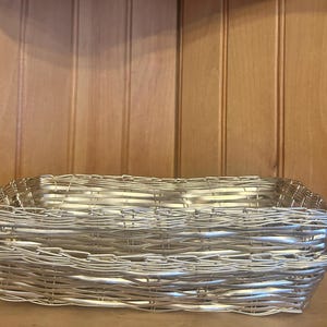 May include: Two silver-colored woven baskets stacked on top of each other. The baskets are rectangular with rounded corners and a tightly woven pattern. The background is a wooden wall.