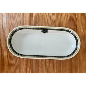 May include: White oval serving dish with a black and gold decorative border. The dish has a black emblem in the center. The dish is on a wooden surface.