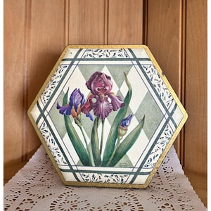 May include: Hexagon shaped box with a gold trim and a floral design. The box features a white background with a green and white checkered pattern. The floral design features three purple irises with green leaves.