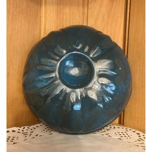 May include: A teal-colored ceramic bowl with a central circular indentation. The bowl has a fluted, petal-like design around the edges. The glossy finish reflects light, highlighting the bowl's shape and color. The bowl is likely for serving or display.
