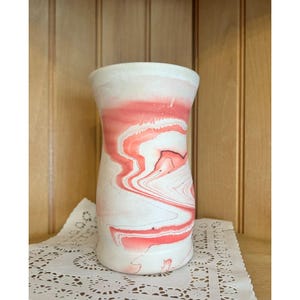 May include: A white ceramic vase with a pink and white marbled design. The vase is about 6 inches tall and has a narrow opening.