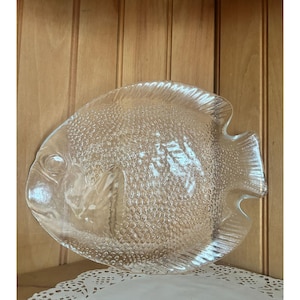 May include: Clear glass fish-shaped serving dish. The dish has a textured surface with a scale-like pattern. The fish's eye and fins are clearly defined. The dish is displayed on a white lace doily against a wooden background.