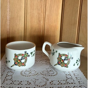 May include: A white ceramic sugar bowl and creamer set with a gold trim. Both pieces feature a Celtic knot design with a sword and shamrocks in green, red, yellow, and blue.