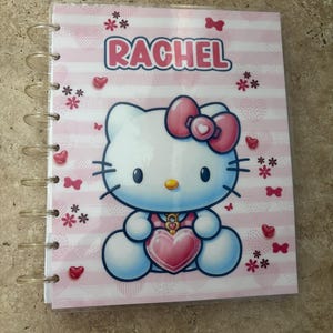 May include: A pink and white spiral-bound notebook featuring a Hello Kitty design. The cover has the name "RACHEL" in pink letters, along with hearts, bows, and flower accents. The notebook is designed with a pink striped background.