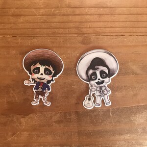 May include: Two stickers featuring cartoon characters in the style of D&iacute;a de Muertos. One is in color, wearing a brown hat and a blue jacket. The other is black and white, holding a guitar and wearing a sombrero.
