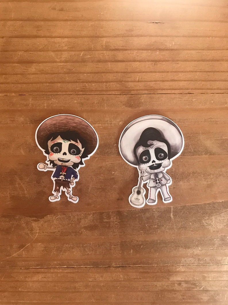May include: Two stickers featuring cartoon characters in the style of D&iacute;a de Muertos. One is in color, wearing a brown hat and a blue jacket. The other is black and white, holding a guitar and wearing a sombrero.