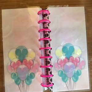 May include: A pink and white planner with a clear plastic cover. The cover has a design of Mickey Mouse balloons in pastel colors.