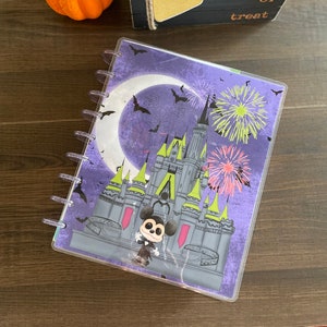 May include: A Halloween-themed planner with a purple cover featuring a castle, fireworks, bats, and a crescent moon. A Mickey Mouse figurine is on the cover. The planner is spiral-bound with clear plastic covers.
