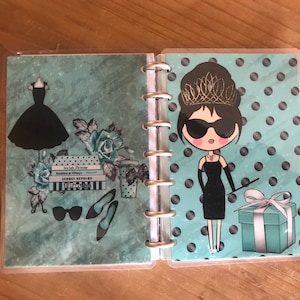 May include: A turquoise blue planner with a black and white polka dot design. The planner features a cartoon illustration of a woman wearing a black dress, sunglasses, and a tiara. The text "Breakfast at Tiffany's" and "Audrey Hepburn" is visible on the planner.