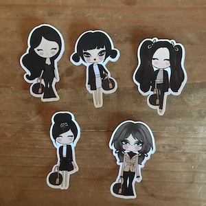 May include: Five illustrated stickers of women with black hair, wearing different outfits and carrying a brown handbag. The stickers are on a wooden surface.