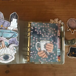 May include: A silver planner with a blue and white starry pattern cover. The planner is open to a page with a sticker of a hand holding a white mug with the text "Magic Spells". The planner has several other stickers on it, including crystals, a moon, an eye, and a book.