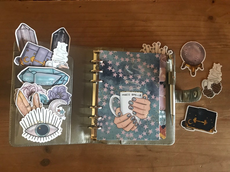 May include: A silver planner with a blue and white starry pattern cover. The planner is open to a page with a sticker of a hand holding a white mug with the text "Magic Spells". The planner has several other stickers on it, including crystals, a moon, an eye, and a book.