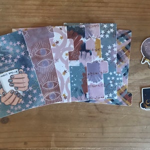 May include: A set of six planner dividers with various designs, including stars, eyes, and abstract patterns. The dividers are printed on a clear, plastic-like material. The dividers are accompanied by three stickers: a crystal ball, a closed book, and a hand holding a mug with the text "Magic Spells".