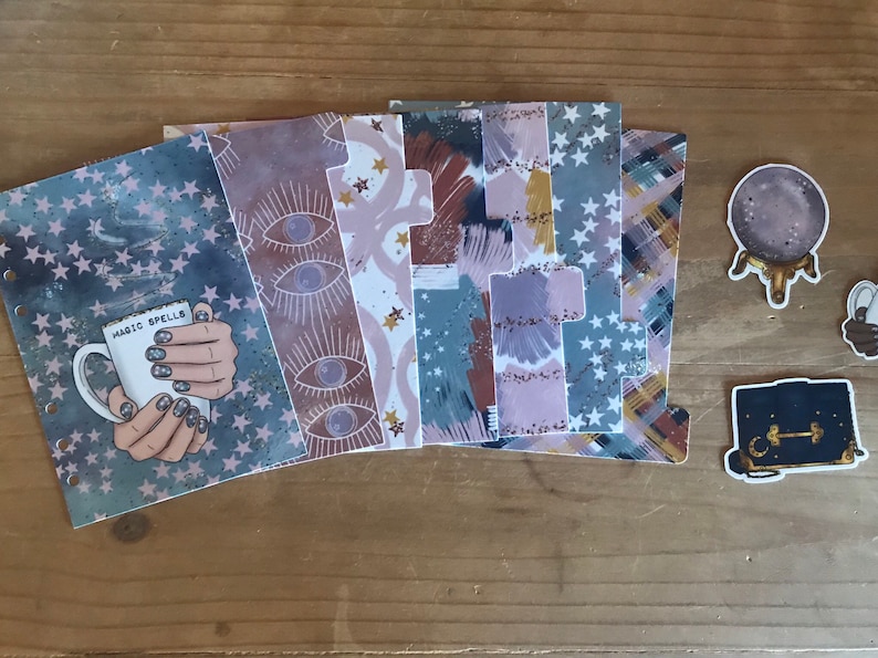 May include: A set of six planner dividers with various designs, including stars, eyes, and abstract patterns. The dividers are printed on a clear, plastic-like material. The dividers are accompanied by three stickers: a crystal ball, a closed book, and a hand holding a mug with the text "Magic Spells".