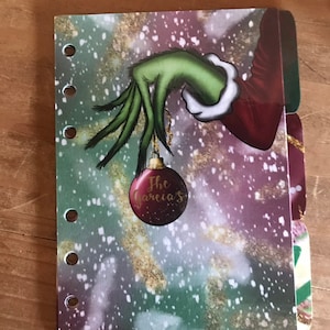 May include: A green Grinch hand holding a red Christmas bauble with the text "The Garcia's" on it. The background is a green, purple, and white snowy design.