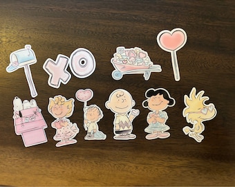 May include: A set of Valentine's Day themed stickers featuring characters from the Peanuts comic strip. The stickers include a mailbox, a wheelbarrow full of hearts, a heart-shaped lollipop, a heart balloon, and various Peanuts characters including Snoopy, Charlie Brown, Lucy, and Sally.