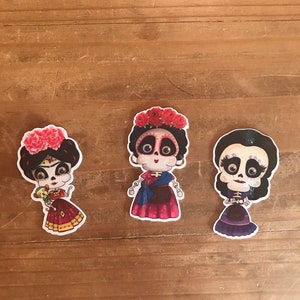May include: Three colorful stickers featuring stylized female figures with sugar skull faces, dressed in vibrant outfits and floral headpieces. The stickers are set against a wooden background, showcasing a Day of the Dead theme.