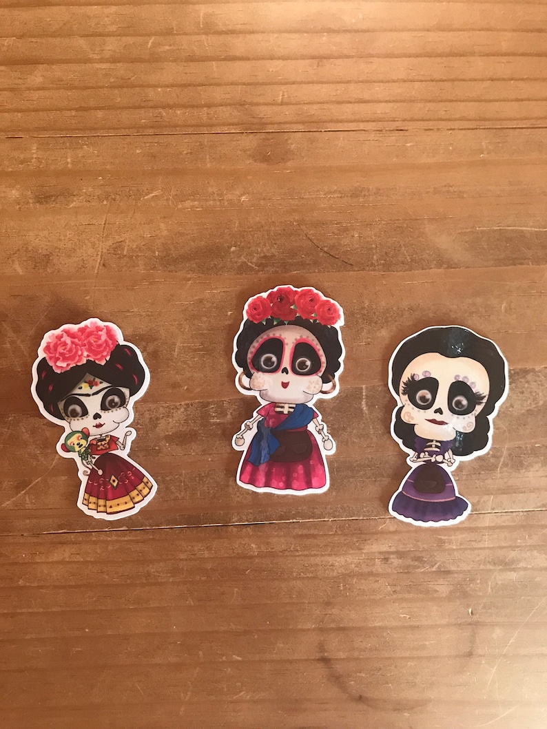 May include: Three colorful stickers featuring stylized female figures with sugar skull faces, dressed in vibrant outfits and floral headpieces. The stickers are set against a wooden background, showcasing a Day of the Dead theme.