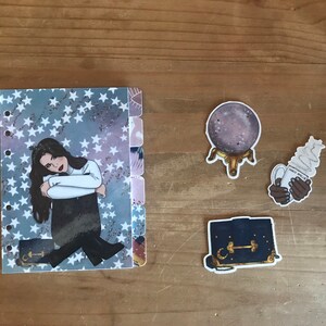 May include: A planner with a blue and white star pattern, a sticker of a woman with long dark hair, a sticker of a crystal ball on a stand, a sticker of a hand holding a cup of coffee with whipped cream, and a sticker of a black purse with a gold clasp.