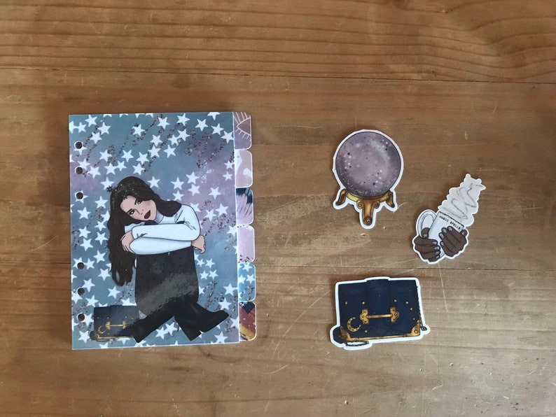 May include: A planner with a blue and white star pattern, a sticker of a woman with long dark hair, a sticker of a crystal ball on a stand, a sticker of a hand holding a cup of coffee with whipped cream, and a sticker of a black purse with a gold clasp.