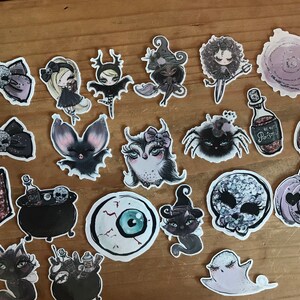 May include: A set of 20 Halloween themed stickers featuring various characters and objects, including a witch, a bat, a spider, a pumpkin, a skull, a cauldron, and a bottle of poison. The stickers are all black and white with glitter accents.