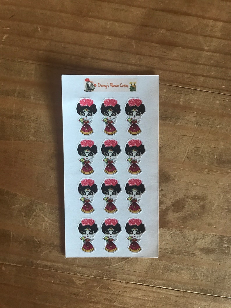 May include: A sheet of stickers featuring a stylized illustration of a woman with a floral headdress and a red dress. The stickers have a Day of the Dead theme. The text "Danny's Planner Cutters" is printed at the top.