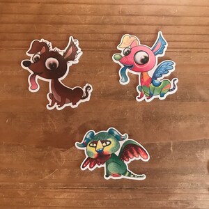 May include: Three colorful cartoon dog stickers with whimsical designs. One is brown with a pink tongue, another is pink with blue wings, and the third is green with red wings. The stickers are on a wooden surface.