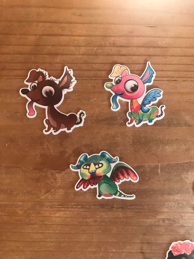 May include: Three colorful cartoon dog stickers with whimsical designs. One is brown with a pink tongue, another is pink with blue wings, and the third is green with red wings. The stickers are on a wooden surface.