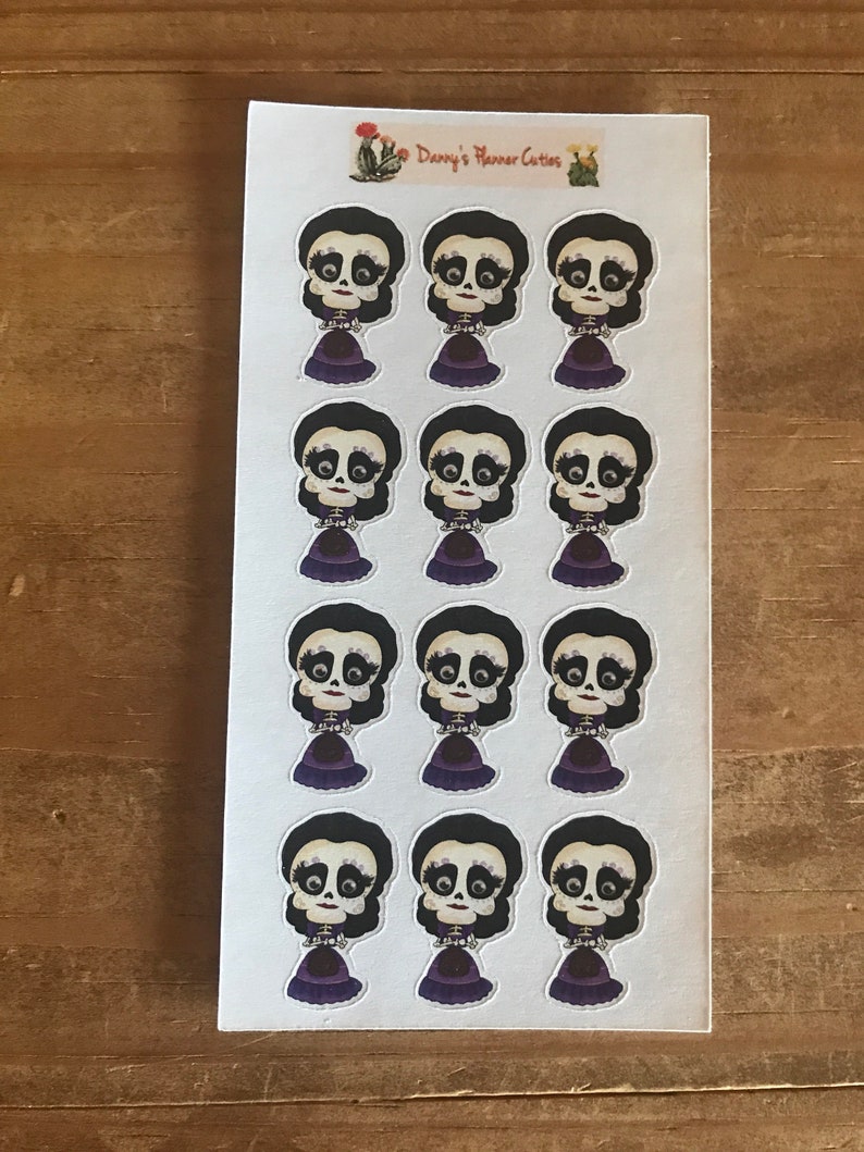 May include: A sheet of stickers featuring a Day of the Dead-themed design. Each sticker depicts a stylized skull with black hair, a purple dress, and a decorative collar. The sheet has the text "Danny's Planner Cuties".