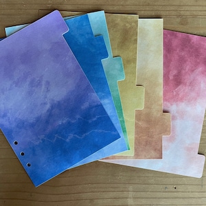 May include: A set of six colorful dividers for a planner or notebook. The dividers are made of paper and have a watercolor-like design. The colors are purple, blue, green, yellow, orange, and pink.