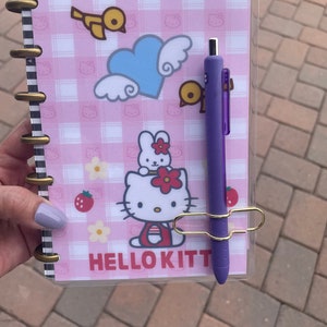 May include: A pink and white checkered planner with a clear cover. The planner features a Hello Kitty design with a bunny and a bird. A purple pen is clipped to the planner with a gold clip.