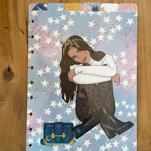 May include: A blue and gold patterned planner cover with a woman sitting on a bed of white stars. The woman is wearing a white sweater, black pants, and black boots. She is holding a blue handbag with a gold clasp and a crescent moon design.