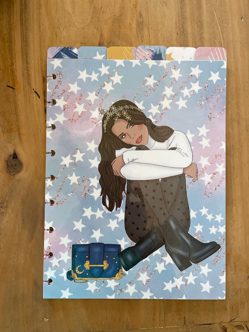 May include: A blue and gold patterned planner cover with a woman sitting on a bed of white stars. The woman is wearing a white sweater, black pants, and black boots. She is holding a blue handbag with a gold clasp and a crescent moon design.