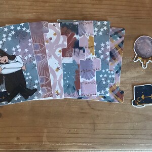 May include: A set of six planner dividers with a celestial theme. The dividers feature a variety of patterns, including stars, eyes, and abstract designs in shades of blue, purple, and brown. The dividers are made of clear plastic and have holes for a planner.