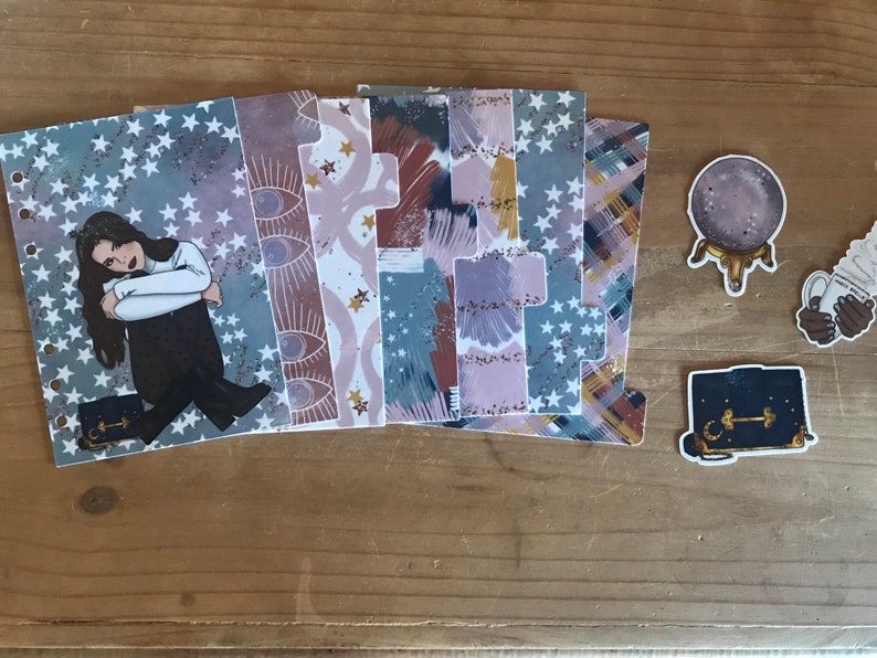 May include: A set of six planner dividers with a celestial theme. The dividers feature a variety of patterns, including stars, eyes, and abstract designs in shades of blue, purple, and brown. The dividers are made of clear plastic and have holes for a planner.