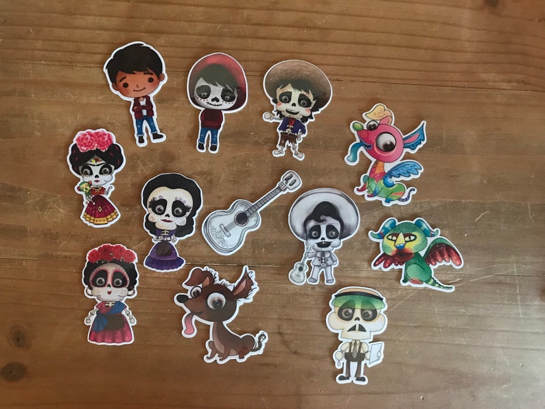 May include: A collection of colorful stickers featuring characters and elements from the animated movie "Coco." The stickers include various character illustrations, a guitar, and fantastical creatures, all with a cartoon style. The stickers are arranged on a wooden surface.