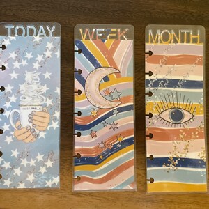 May include: Three clear plastic planner dividers with colorful designs. The first divider has a blue background with white stars and a coffee cup with the text "Magic Spells". The second divider has a rainbow striped background with a crescent moon and stars. The third divider has a rainbow striped background with a blue eye. The dividers are labeled "TODAY", "WEEK", and "MONTH".