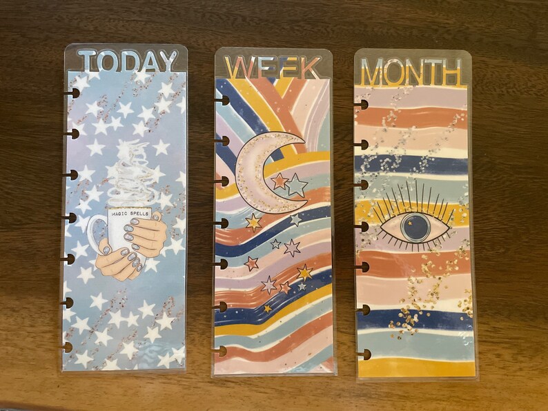 May include: Three clear plastic planner dividers with colorful designs. The first divider has a blue background with white stars and a coffee cup with the text "Magic Spells". The second divider has a rainbow striped background with a crescent moon and stars. The third divider has a rainbow striped background with a blue eye. The dividers are labeled "TODAY", "WEEK", and "MONTH".