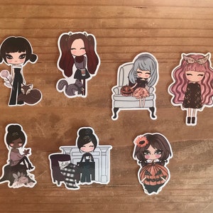 May include: A collection of seven stickers featuring stylized illustrations of girls in various autumnal settings. The stickers showcase girls with different hairstyles and outfits, some holding pumpkins, sitting in chairs, or with animals. The color palette includes browns, blacks, and pinks.